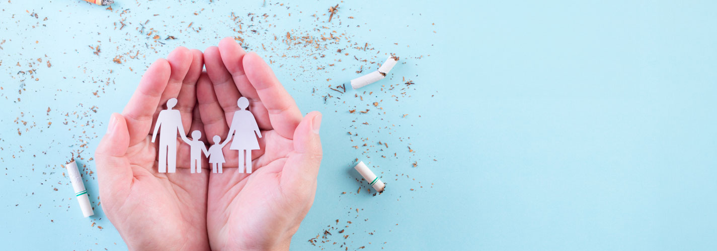 A set of hands holds a paper cut out of a family, protecting them from the cigarettes on the light blue background.
