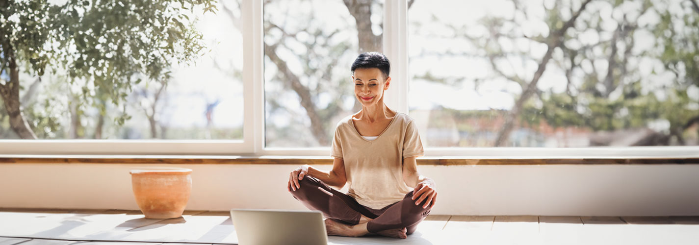 Mature woman meditating near laptop in front of large sunny windows