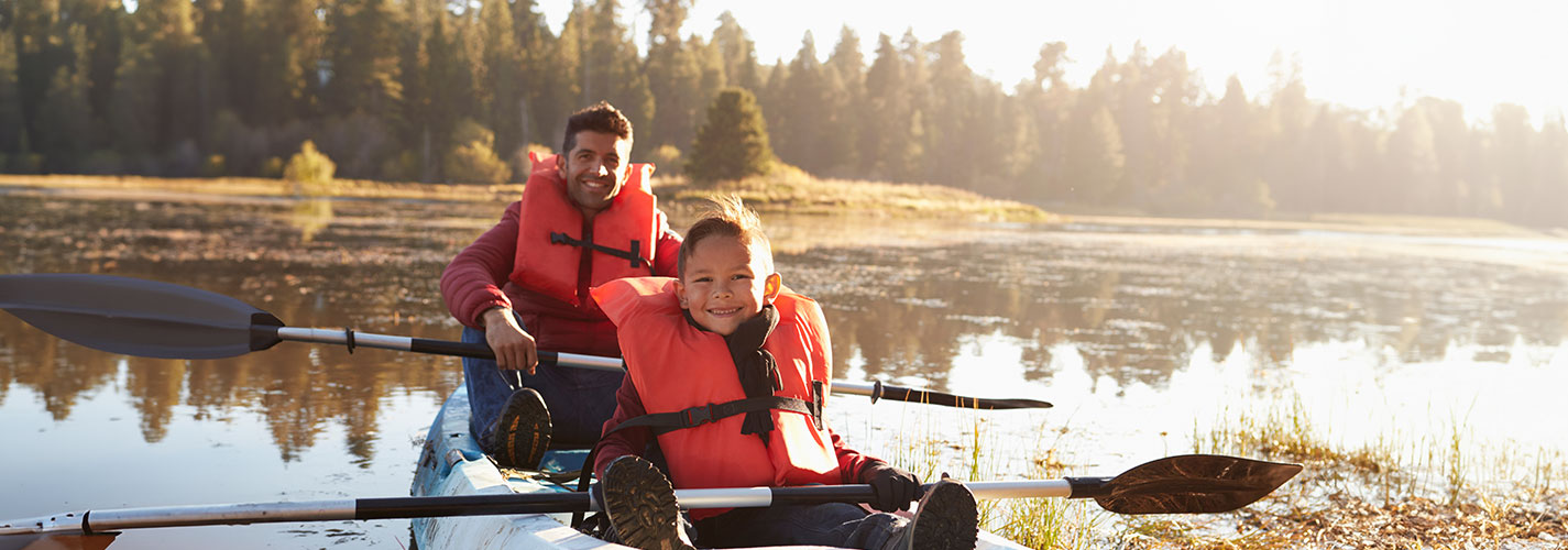 A father and son wearing life jackets, as they kayak on the lake together.
