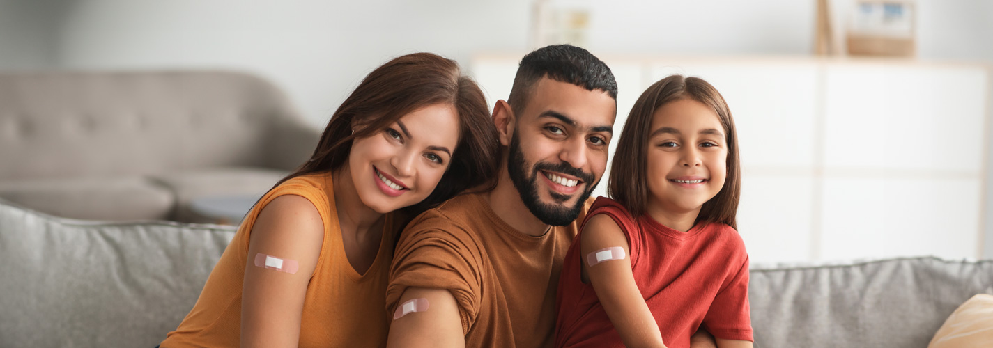 A smiling family of a mother, father, and daughter sit on a grey couch, showing their bandage after their vaccines.