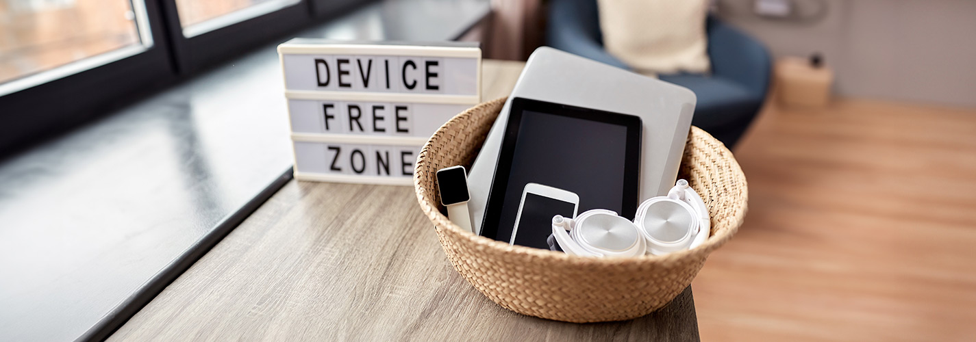 A wicker basket filled with tablets, a smart phone, watch and headphones. Next to it is a lightbox which reads "Device Free Zone."