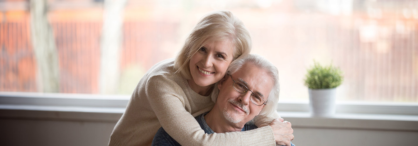 A smiling woman stands behind her husband, embracing him in a hug.