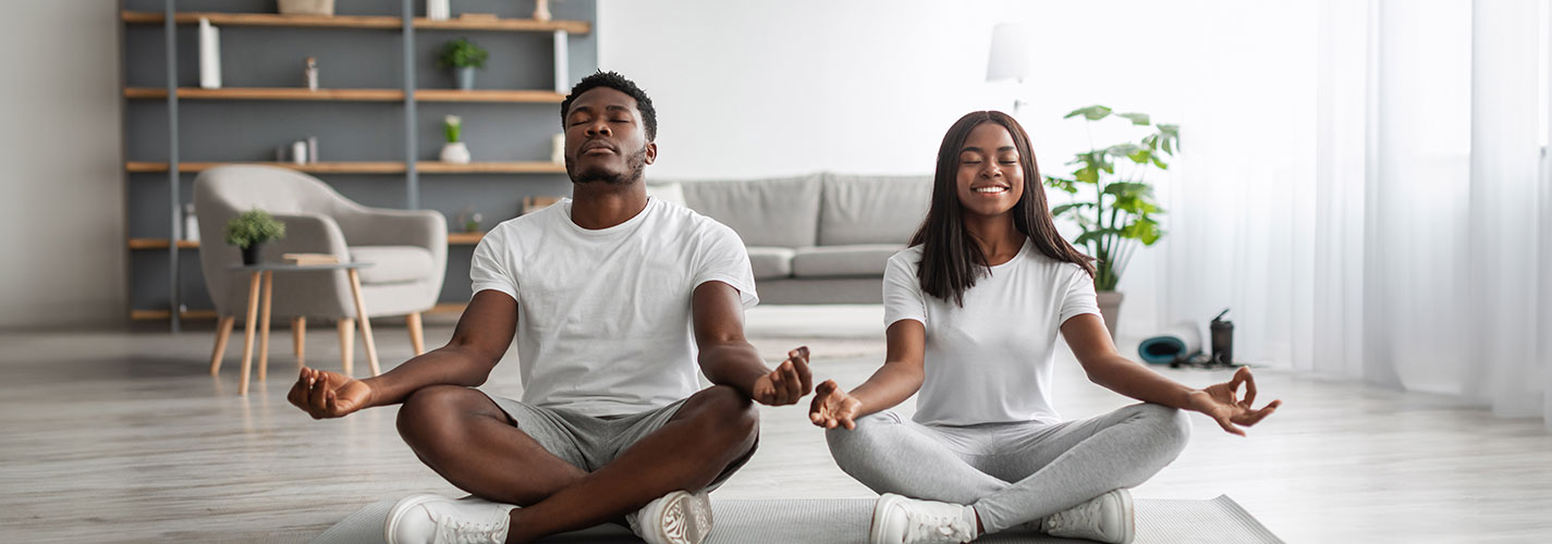 An African American man and woman sit cross-legged on a grey yoga mat and meditate together.