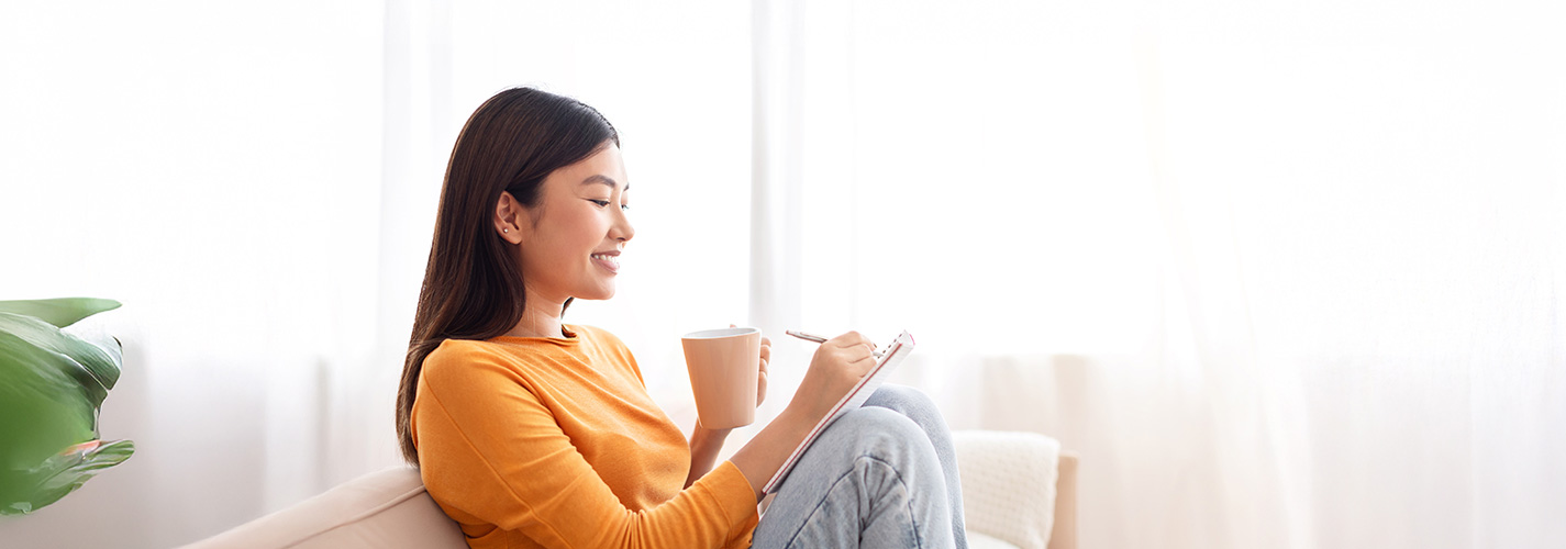 A young woman happily sits on her couch and writes in her journal, in a bright airy living room.