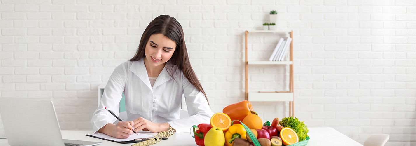 A woman sits at a desk with a variety of fresh fruit and vegetables next to her as she writes in a notebook.