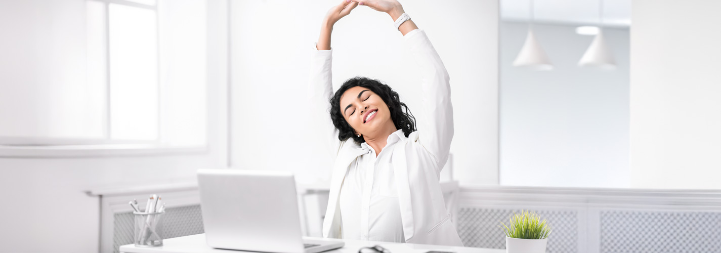 A young woman stretches her hands above her head, taking a break from working on her laptop.